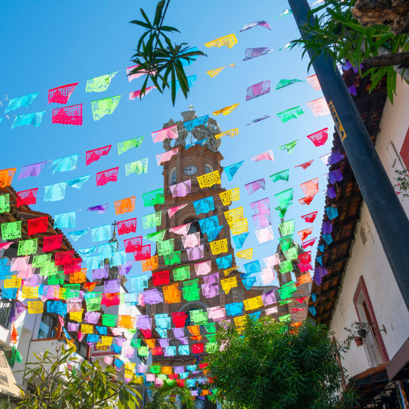 View from the street below of the The Church of Our Lady of Guadalupe with colorful flags strung across the street on a sunny morning in Puerto Vallarta, Mexico