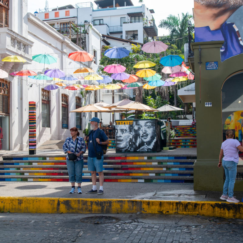 A couple stands near a colorful umbrella display near restaurants and shops in the popular Zona Romantica in Puerto Vallarta