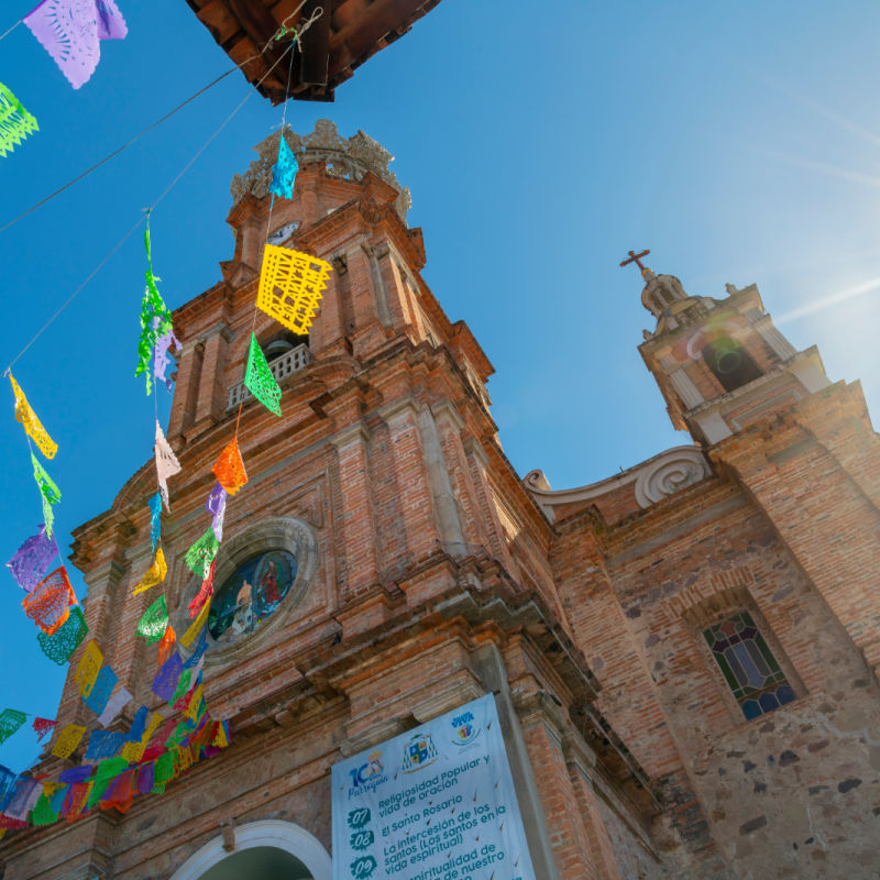 View from the street below of the The Church of Our Lady of Guadalupe with colorful flags strung across the street on a sunny morning in Puerto Vallarta, Mex