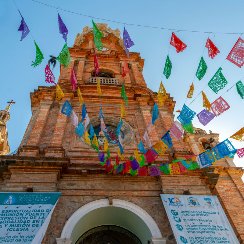 View from the street below of the The Church of Our Lady of Guadalupe with colorful flags strung across the street on a sunny morning in Puerto Vallarta, Mexico